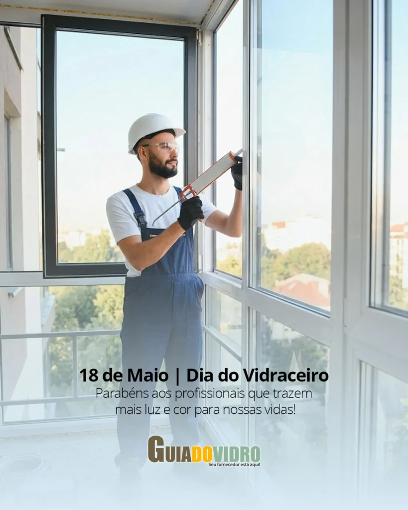 Imagem colorida em plano médio de um vidraceiro com barba, usando capacete de segurança branco, óculos protetores e macacão jeans azul sobre camiseta branca. Ele segura uma pistola de calafetagem laranja e aplica um cordão de selante na junção de um grande painel de vidro em uma sacada de alumínio e vidro. A janela se estende do chão ao teto e reflete a luz do dia e uma paisagem urbana arborizada ao fundo. No canto superior esquerdo, sobrepostos em branco, estão o título "18 de Maio | Dia do Vidraceiro" e a mensagem de parabéns. No chão da sacada, há um rolo de cabo e uma caixa de ferramentas.